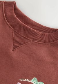 Burgunder sweatshirt med ribbet rund hals, en tekstureret trekantdetalje ved kraven og broderet "SEASONS" med en grøn sløjfe accent.