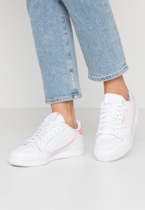 Dolní končetiny v světle modrých džínách a bílých teniskách Adidas s růžovými akcenty, stojící na světlé podlaze proti jednobarevnému pozadí.