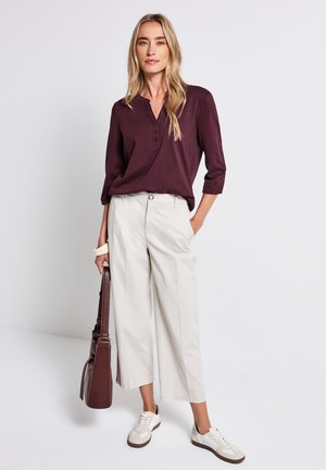 Chemise henley à manches longues bourgogne, pantalon large crème et baskets blanches. Sac à main marron porté en bandoulière avec des accents de texture légère.