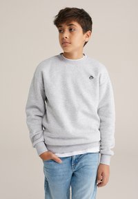 Világosszürke pulóver kerek nyakkivágással és bordázott mandzsettákkal, kis sötét logóval a mellkasán. Világoskék farmerrel párosítva.