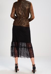 Top metallizzato marrone con vita stretta, abbinato a una gonna nera in pizzo a strati. Scarpe nere con tacco alto completano l'outfit.