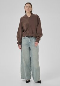 Brown cropped zip-up sweatshirt with wide sleeves paired with light blue wide-leg jeans and black pointed-toe shoes. Plain background.
