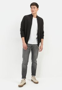 Cardigan zippé noir sur un t-shirt blanc, associé à un jean gris et des baskets beiges. Le modèle se tient contre un fond blanc.