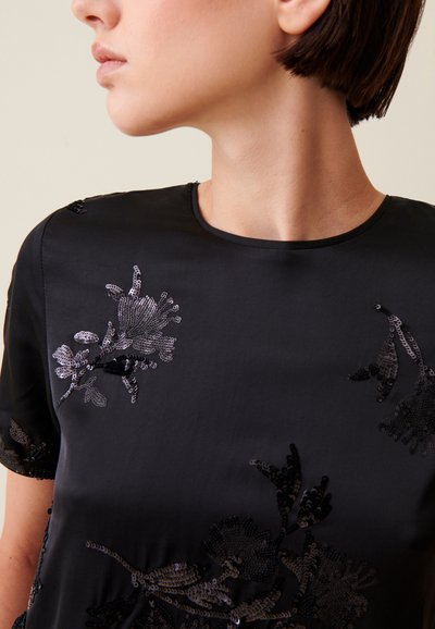 Donna con capelli corti e scuri che indossa una maglia nera con ricami di paillettes floreali sul davanti e le maniche.