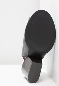 Semelle d'une chaussure à talon en cuir noir avec fond en caoutchouc texturé et talon carré, inclinée contre un fond de sol et de mur blancs.