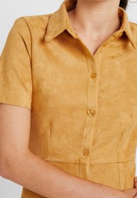 Camicia a maniche corte, con colletto, in camoscio giallo senape, con chiusura frontale a bottoni e texture liscia. I dettagli dei bottoni sono ben visibili.
