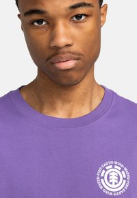 Lilla bomulds T-shirt med rund halsudskæring og korte ærmer, med et hvidt cirkulært grafik på brystet, der har tekst og logo detaljer.