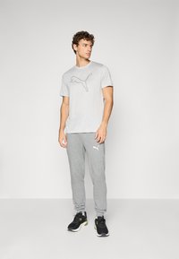 Grå t-shirt med sort Puma-logo, parret med lysegrå sweatpants med et hvidt logo. Sorte træningssko med grønne accenter.