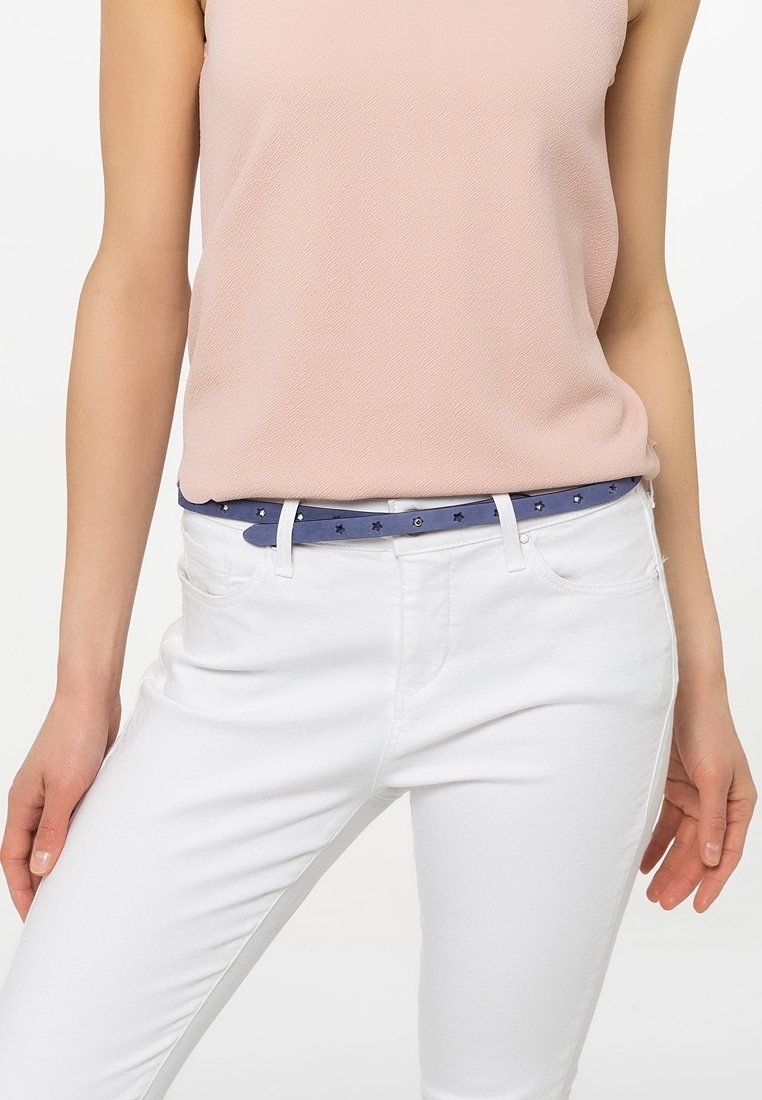 Femme portant un haut sans manches rose clair, un pantalon blanc et une ceinture bleue avec des découpes en forme d'étoile autour de la taille.
