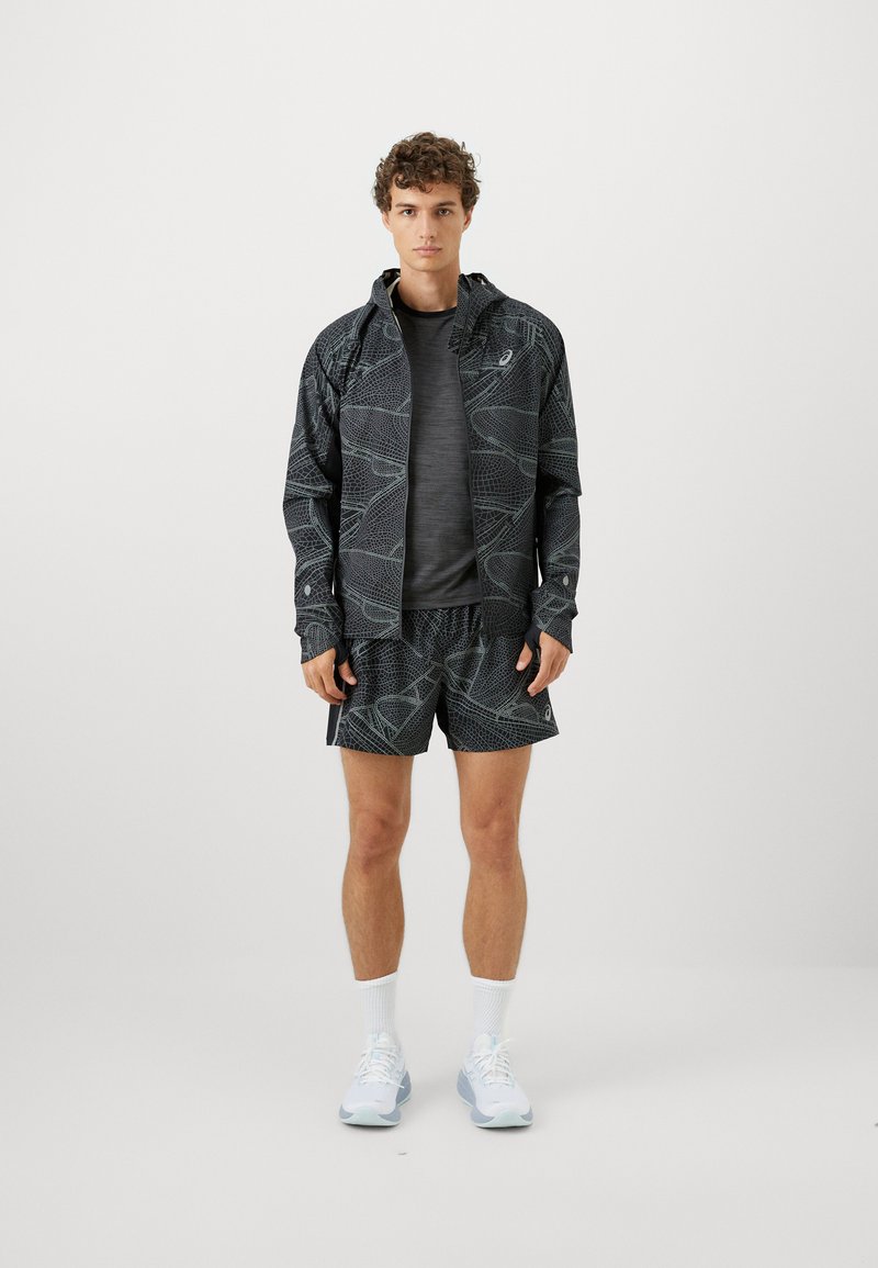 Chaqueta deportiva negra con patrón geométrico, combinada con pantalones cortos negros lisos. Modelo lleva zapatillas de correr azul claro y calcetines blancos.