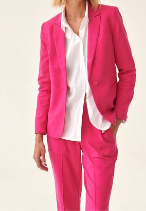 Frau trägt einen knallpinken Anzug mit maßgeschneidertem Blazer und passender Hose über einem weißen Hemd und steht mit einer Hand in der Tasche.