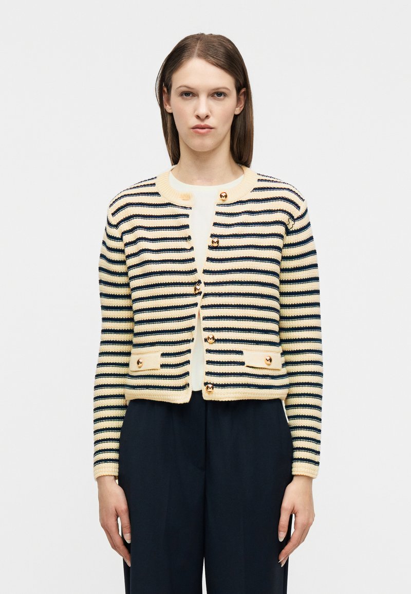 Γυναίκα που φοράει ριγέ ζακέτα σε εκρού και ναυτικό μπλε με χρυσά κουμπιά, λευκή μπλούζα από κάτω και σκούρο ναυτικό μπλε παντελόνι με φαρδιά μπατζάκια.