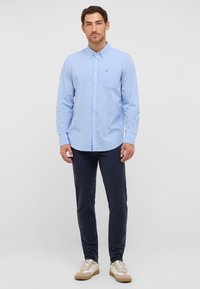 Blau-weiß kariertes Hemd mit Button-down-Kragen, langen Ärmeln und einer Brusttasche, kombiniert mit dunkelblauen Hosen und beigen Turnschuhen.