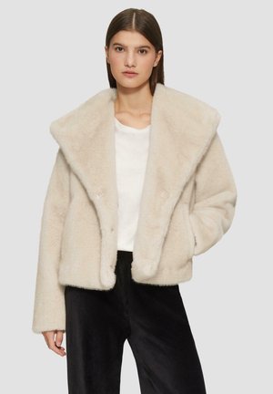Veste en fausse fourrure couleur crème avec un large col, une fermeture à boutons et des poches latérales. Associée à une chemise blanche et un pantalon noir.
