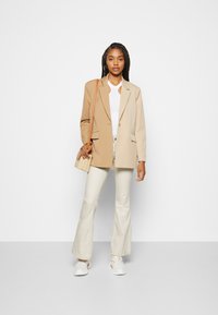 Blazer bicolor bege, blusa branca com recorte, jeans cream com boca de sino, sapatilhas brancas, segurando uma mala texturizada em ton de bege.