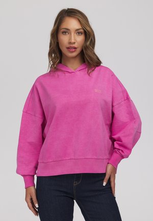 Kobieta z falującymi jasnobrązowymi włosami, ubrana w jaskrawy różowy oversize'owy kaptur i ciemnoniebieskie dżinsy, stojąca na jednolitym szarym tle.