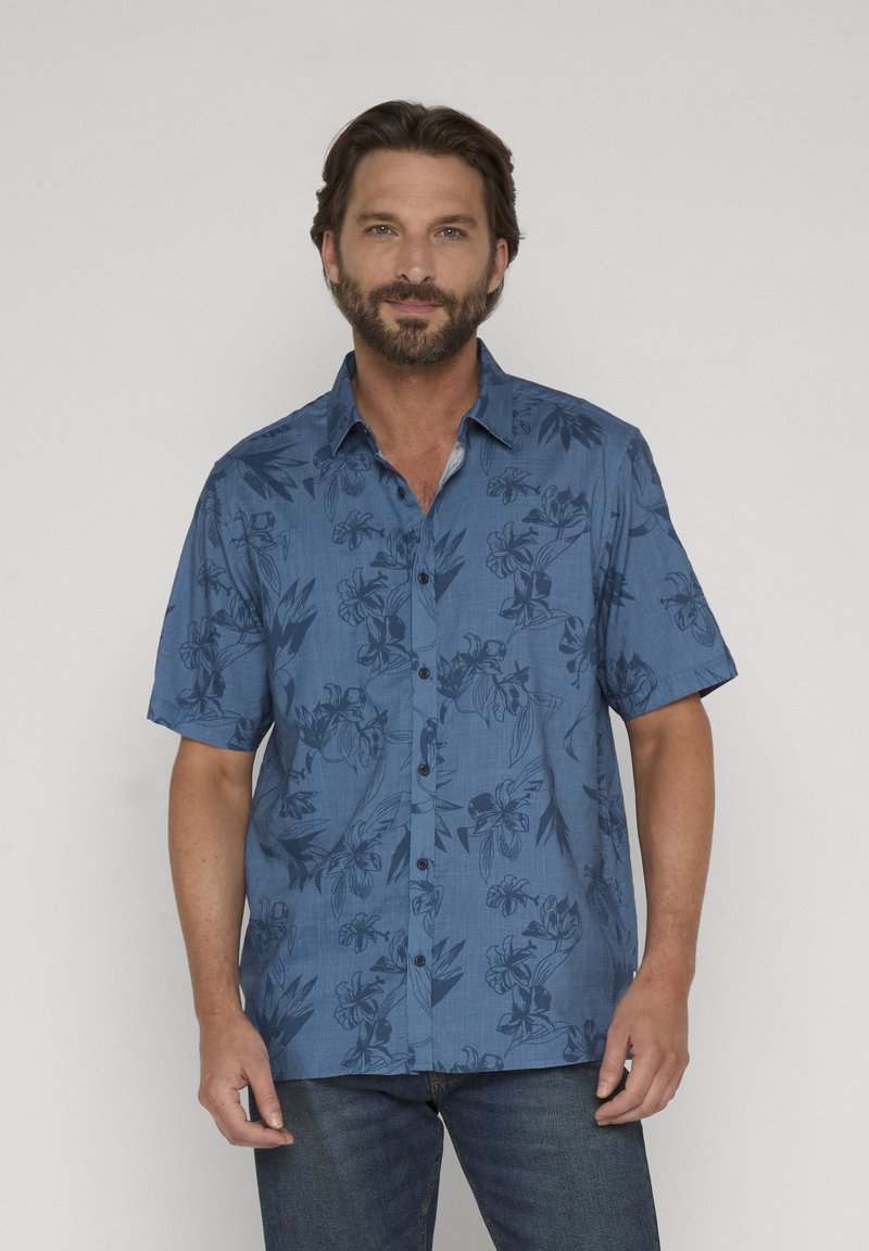 Homme portant une chemise à manches courtes bleue à boutons avec un imprimé floral foncé et un jean bleu foncé, debout devant un fond gris uni.