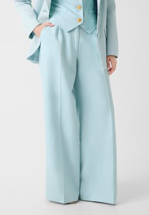 Kobieta ubrana w jasnoniebieskie szerokie spodnie i dopasowaną marynarkę z złotymi guzikami, stojąca z jedną ręką w kieszeni.