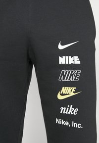 Pantalones deportivos negros de algodón con múltiples logotipos de Nike en varios colores y fuentes impresos en el lateral, con una textura suave.