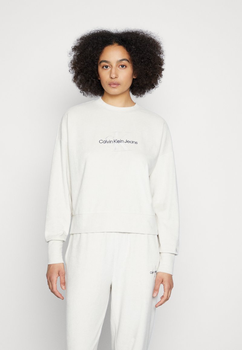 Světle béžová mikina s dlouhými rukávy, žebrovanými manžetami a zkráceným lemem. Na hrudníku se nachází logo "Calvin Klein Jeans" v tmavě šedé barvě.