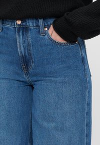 Blugi din denim de un albastru deschis, cu talie înaltă, având accesorii din cupru, buzunare frontale și un design drept. Purtați împreună cu un pulover negru tricotat.