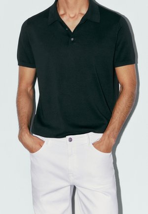 Homme portant un polo noir à manches courtes rentré dans un pantalon blanc, mains posées dans les poches avant, sur un fond clair uni.