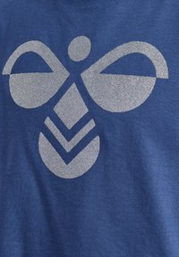 Blaues T-Shirt mit einem großen, glitzernden silbernen Grafikdesign mit abgerundeten Formen und Chevron-Mustern in der Mitte.