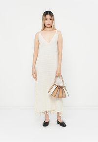 O rochie lungă, de culoare crem, cu decolteu în V, realizată dintr-un material texturat, are franjuri la tiv. Modelul ține o poșetă multicoloră.