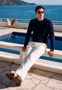 Homme portant des lunettes de soleil, un pull bleu marine, un pantalon blanc et des chaussures marron assis sur une rambarde blanche près d'une piscine avec l'océan et des collines en arrière-plan.