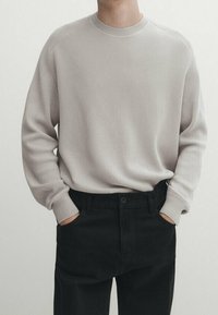 Pull en maille côtelée beige clair avec un col rond, associé à un pantalon noir. Le pull a des manches longues et une coupe décontractée.