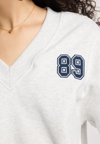 Light gray sweatshirt featuring a V-neck design and navy blue embroidered number "89" on the left chest area. Soft, textured fabric.
