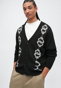 Jeune homme avec des cheveux tressés portant un cardigan noir boutonné avec des motifs de serpent et de dés blancs sur une chemise blanche et un pantalon beige.