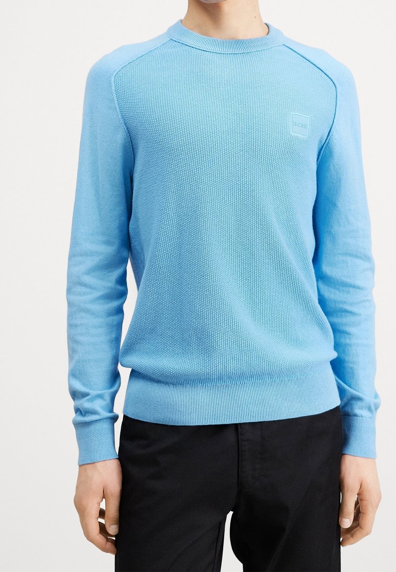 Hellblauer Strickpullover mit Rundhalsausschnitt, gerippten Bündchen und Saum, aus strukturiertem Stoff und mit einem kleinen Markenlogo auf der Brust.