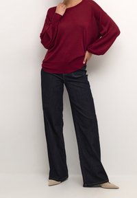 Pull en tricot bordeaux à coupe ample et manches larges, associé à un jean à jambes larges bleu foncé et des chaussures beige à bout pointu.