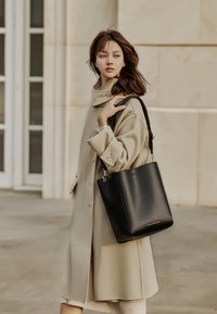 Zwarte leren tas met een gladde textuur en gouden hardware, vastgehouden door een persoon in een beige jas, tegen een effen achtergrond.