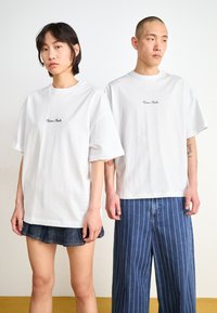 Hvite kortermede t-skjorter med rund hals og liten svart logo, båret av to personer, kombinert med denimbukser i ulike stiler.