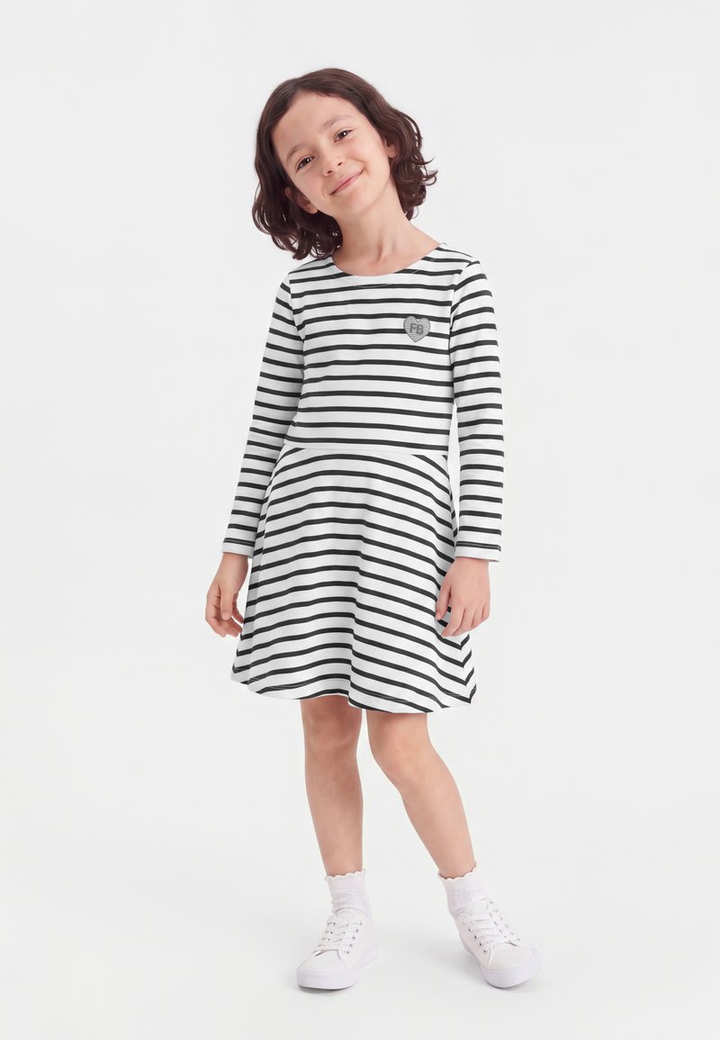 Jeune fille aux cheveux courts et foncés, portant une robe blanche à rayures noires horizontales, des chaussettes blanches et des baskets blanches, debout et souriante.