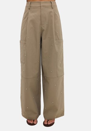 Pantalon beige taille haute à jambes larges avec poches plaquées sur les côtés, décoré de petits clous métalliques, porté avec des sandales ouvertes foncées.