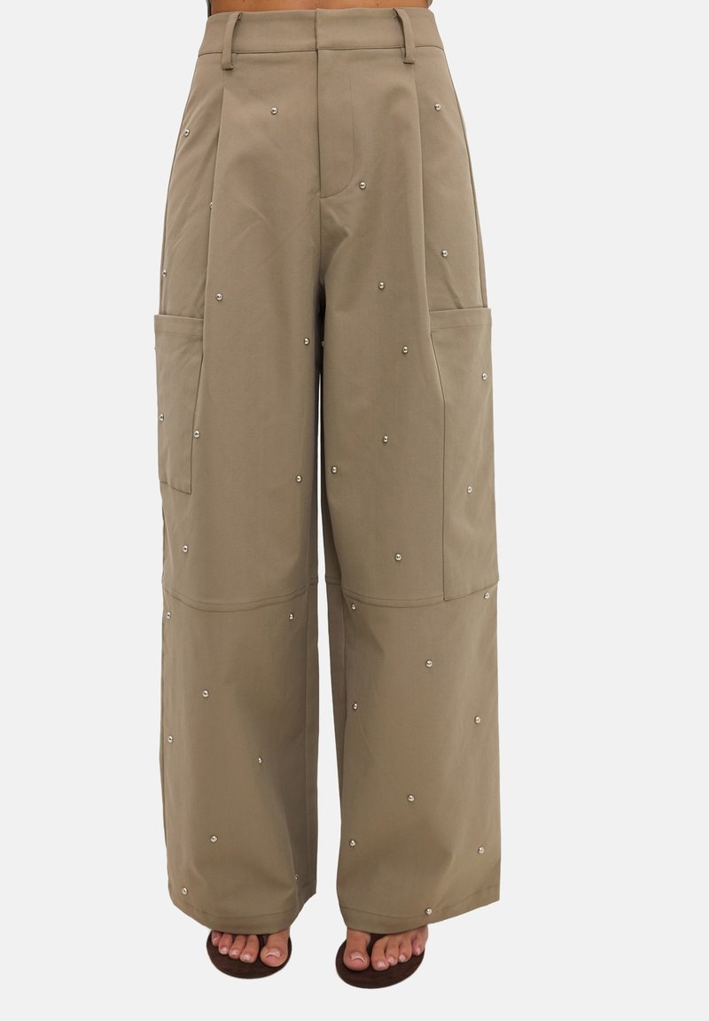 Pantalon beige taille haute à jambes larges avec poches plaquées sur les côtés, décoré de petits clous métalliques, porté avec des sandales ouvertes foncées.