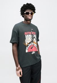 Μαύρο γραφικό μπλουζάκι με τη φράση «AUTHENTIC JORDAN», που φιλοξενεί σχέδιο με παίκτη μπάσκετ και γήινη σφαίρα. Φορεμένο με γυαλιά ηλίου και σκούρο παντελόνι.