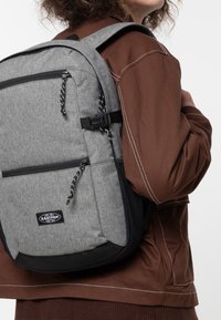 Person wearing a gray Eastpak backpack with black zippers and adjustable straps, dressed in a brown jacket with white stitching.