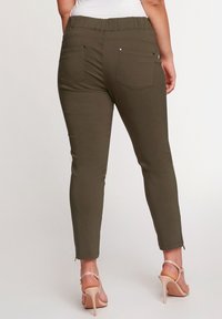 Olivově zelené, těsně přiléhavé kalhoty se pružným pasem, dvěma zadními kapsami a zipy na kotnících. Materiál se zdá být hladký s mírným leskem.