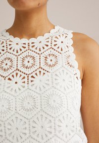 Vit virkade topp med ett hexagonmönster och blommotiv, vågig halsringning och texturerade detaljer genom hela tyget.
