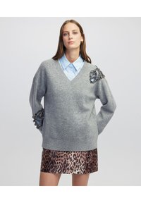 Grijze gebreide trui met een V-hals en decoratieve versiering op de schouder. Gecombineerd met een lichtblauw overhemd met kraag en een rok met luipaardprint.