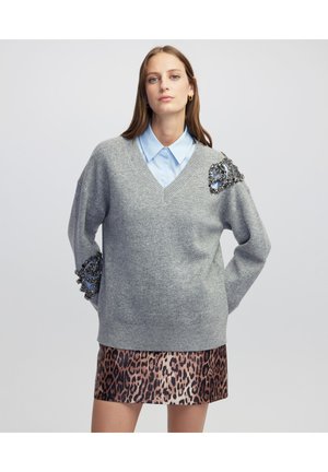 Pull en tricot gris avec un col en V et un embellissement décoratif sur l'épaule. Associé à une chemise col bleu clair et une jupe imprimé léopard.