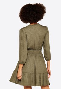 Olivenfarbenes Kleid mit dreiviertel Puffärmeln, gerafftem Taillenbund und ausgestelltem Rock. Der strukturierte Stoff weist ein dezentes Diamantmuster auf.