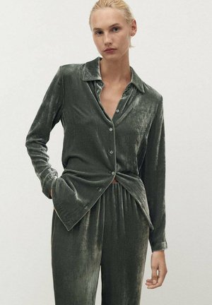 Completo loungewear in velluto di verde tenue. Presenta una camicia con colletto e bottoni e pantaloni con vita elastica, enfatizzando una texture morbida e una lucentezza delicata.