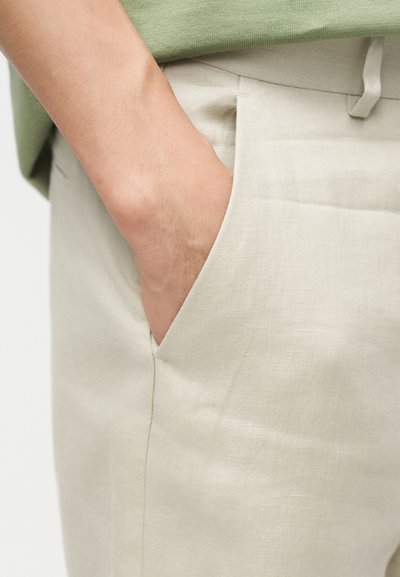 Χέρι τοποθετημένο μέσα στην εμπρόσθια τσέπη μπεζ παντελονιού λινό, συνδυασμένο με πράσινο πουκάμισο.