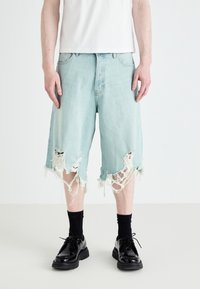 Șorturi din denim de un albastru deschis, cu deschideri mari și desfăcute la margine, purtate cu pantofi negri și șosete negre. Design simplu, croială relaxată.