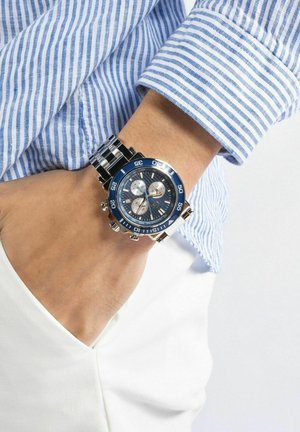 Chronograaf horloge met een blauwe bezel, een zilveren roestvrijstalen band en gedetailleerde subdials. Draagt op een pols met een gestreept blauw overhemd.